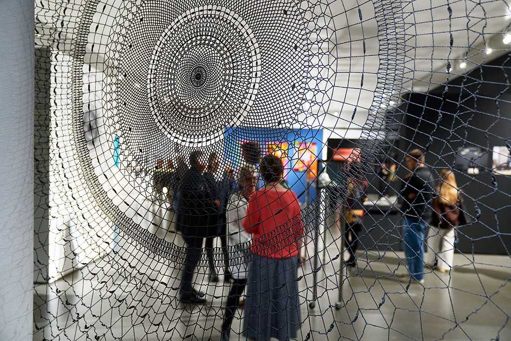 Blick durch ein kunstvoll gewebtes Netz auf einen Raum mit Ausstellung und Menschen