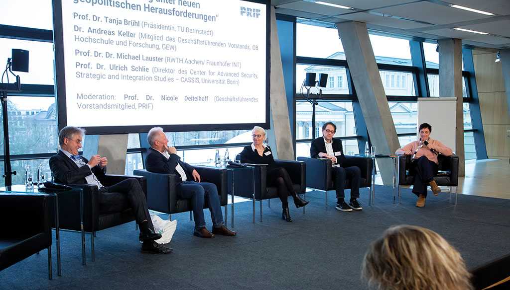 Podium, auf dem drei Männer und zwei Frauen sitzen, eine Frau spricht in ein Mikrofon.