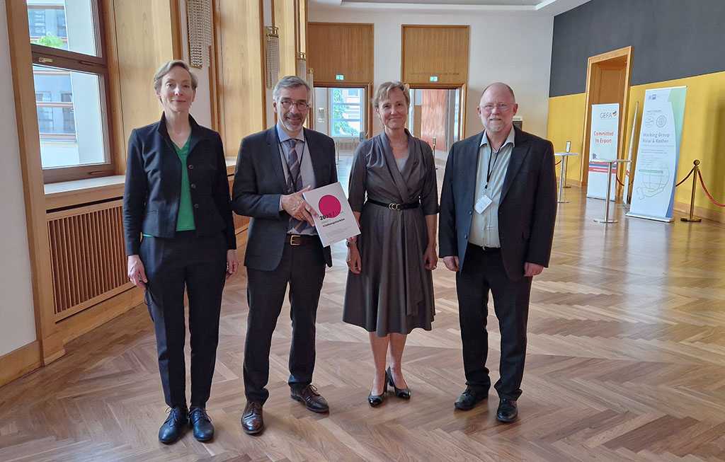 Eine Gruppe von Personen steht in einem großen Saal, ein Mann in der Mitte hält das Friedensgutachten 2025 in den Händen.