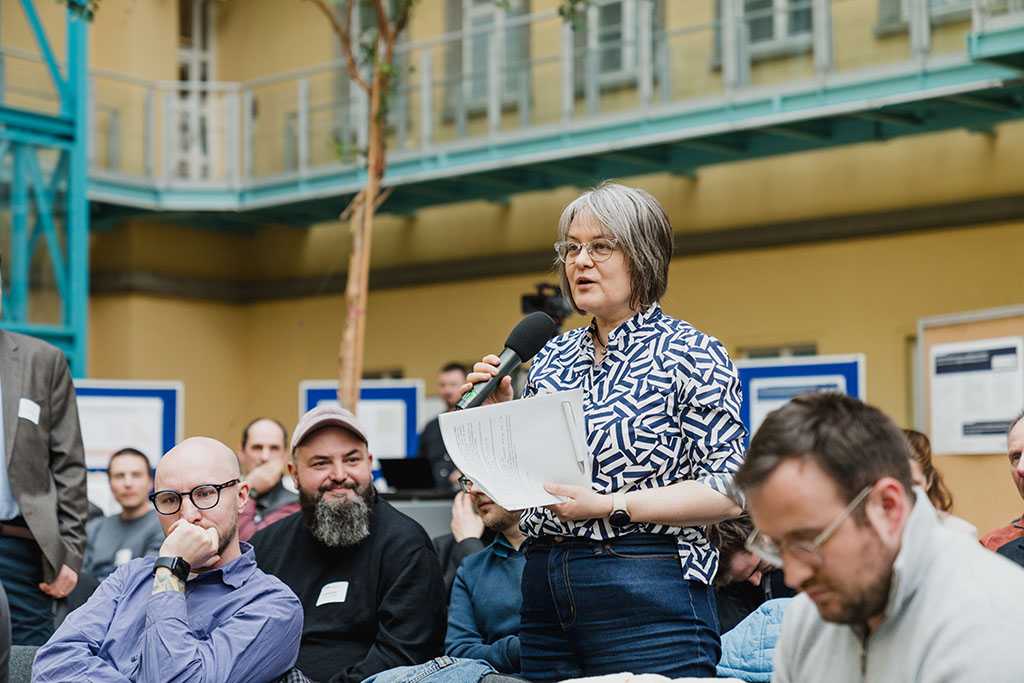 Gefüllter Raum mit sitzenden Menschen, eine Frau steht, mit einem Blatt Papier und einem Mikrofon in der Hand