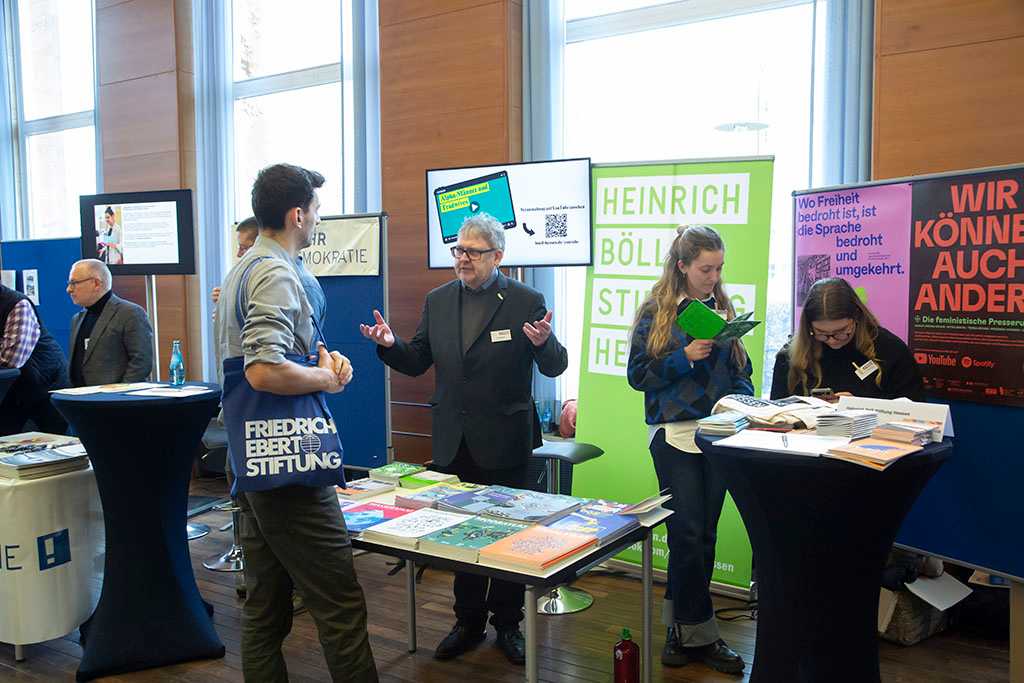 Stand mit ausgelegten Büchern und Plakaten, Menschen unterhalten sich und betrachten die Bücher